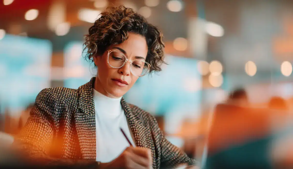 Professional woman with glasses writing in a notebook while working on a laptop in a modern, softly blurred office setting.