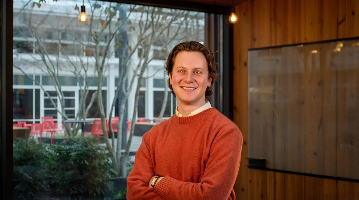 Harry Morgan smiling with arms crossed, wearing an orange sweater indoors