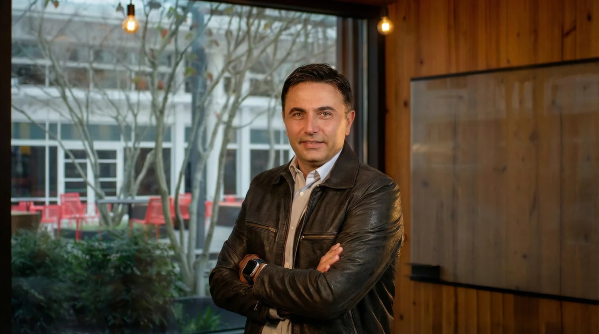 Mark Ghazai in a leather jacket, arms crossed, by a window