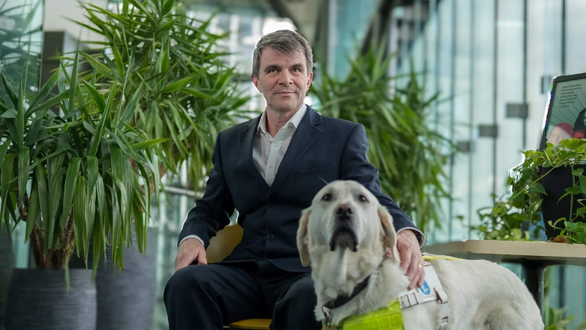 Robin Christopherson seated in a dark suit beside his guide dog